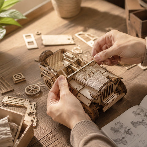 Adult hands assembling a vintage off-road vehicle 3D wooden model kit on a wooden workbench.
